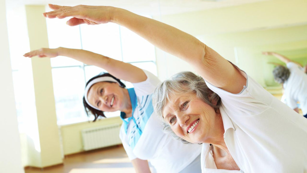 A picture of two individuals smiling and leaning with an arm overhead to stretch the sides of their bodies.